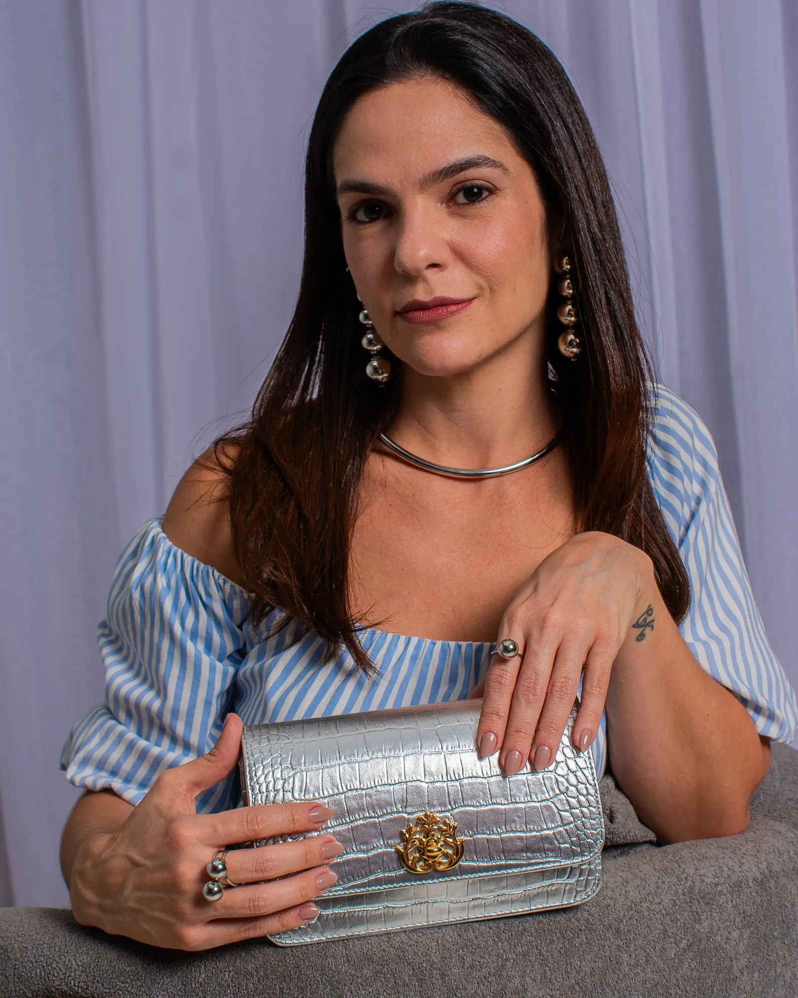 Modelo sentada segurando clutch prata de textura croco, com colar e brincos